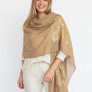 May include: A tan-colored shawl with gold paisley designs. The shawl is draped over the shoulders and features intricate patterns. The fabric appears soft and lightweight, suitable for layering.
