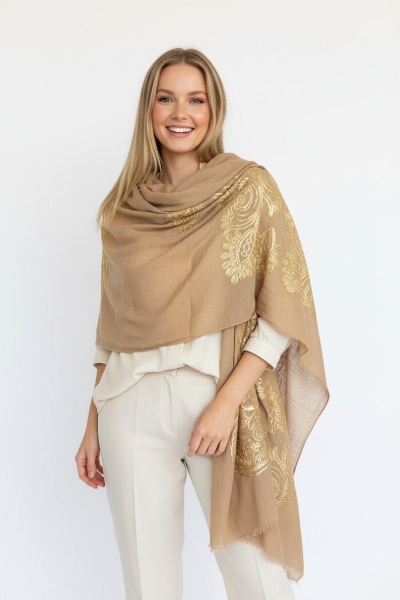 May include: A tan-colored shawl with gold paisley designs. The shawl is draped over the shoulders and features intricate patterns. The fabric appears soft and lightweight, suitable for layering.