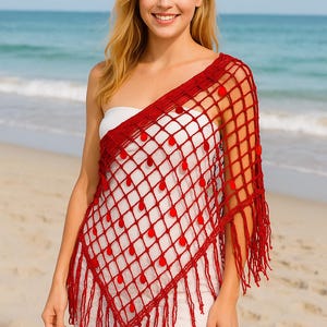 Puede incluir: Un chal rojo de ganchillo con un patrón de diamantes y flecos. El chal presenta pompones rojos y se usa sobre un vestido blanco. El fondo muestra una playa con el océano a lo lejos.