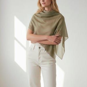 May include: A woman wearing a sage green shawl and off-white jeans. The lightweight shawl drapes over the shoulders and arms. The jeans are high-waisted with a straight leg. The background is a plain white wall.