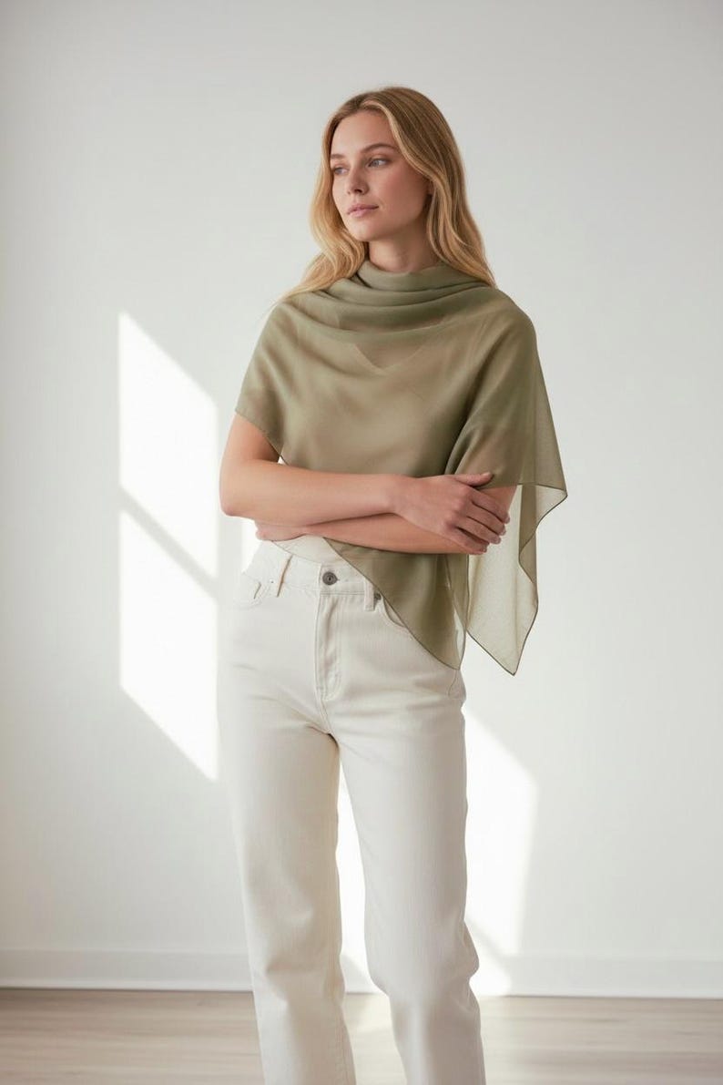 May include: A woman wearing a sage green shawl and off-white jeans. The lightweight shawl drapes over the shoulders and arms. The jeans are high-waisted with a straight leg. The background is a plain white wall.