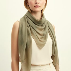 May include: A light olive green scarf draped around the neck. The scarf is made of a sheer, lightweight fabric. The model is wearing a cream-colored top and pants. The background is a solid off-white color.