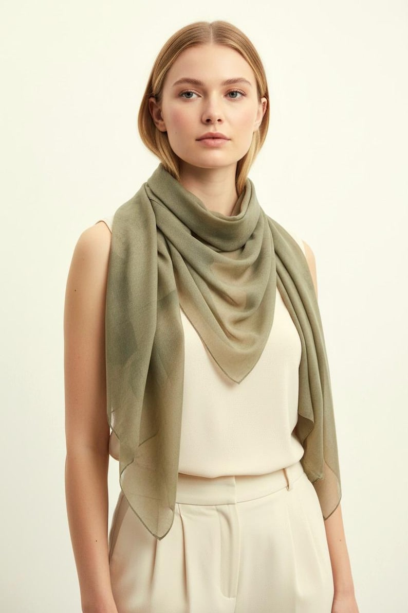 May include: A light olive green scarf draped around the neck. The scarf is made of a sheer, lightweight fabric. The model is wearing a cream-colored top and pants. The background is a solid off-white color.