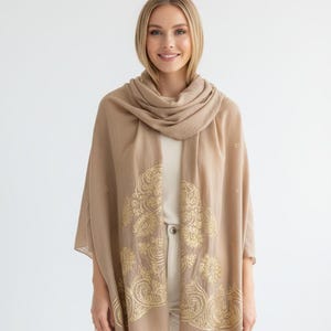 May include: A tan-colored scarf with a gold floral design. The scarf is draped over the shoulders and has a soft, flowing appearance. The gold pattern is intricate and detailed, adding a touch of elegance.