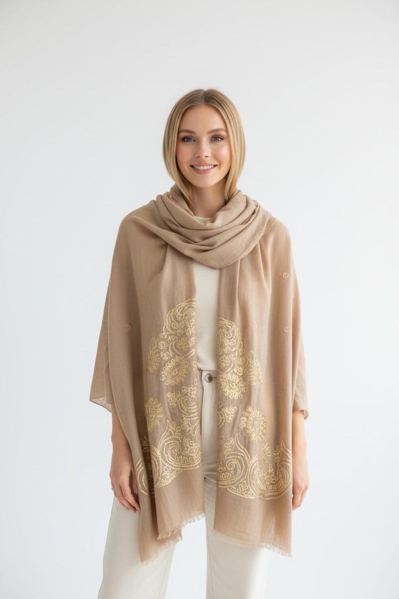 May include: A tan-colored scarf with a gold floral design. The scarf is draped over the shoulders and has a soft, flowing appearance. The gold pattern is intricate and detailed, adding a touch of elegance.