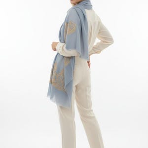 May include: A light blue scarf with a gold paisley design is draped over a cream-colored blouse and pants. The scarf is made of a lightweight fabric and has a fringed edge. The outfit is simple and elegant.