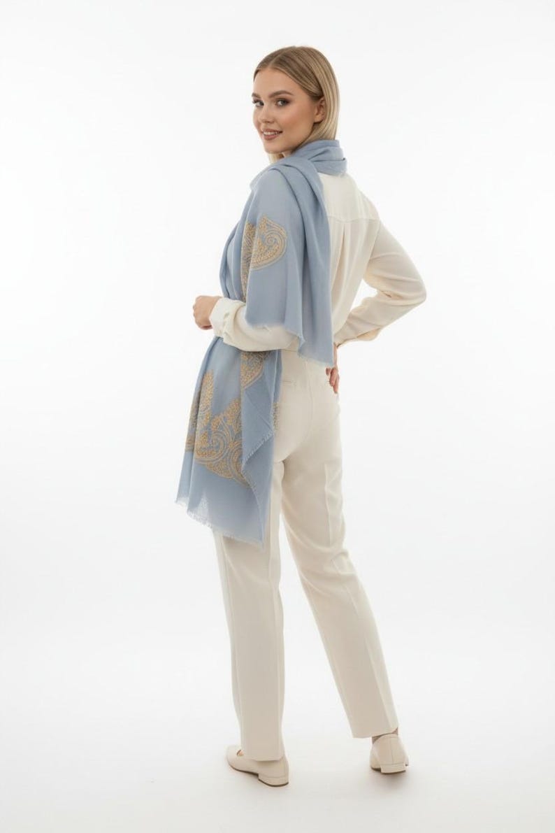 May include: A light blue scarf with a gold paisley design is draped over a cream-colored blouse and pants. The scarf is made of a lightweight fabric and has a fringed edge. The outfit is simple and elegant.