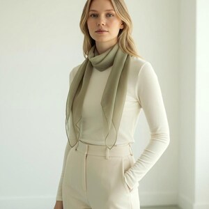 May include: A woman wearing a light green scarf, a white long-sleeve shirt, and cream-colored pants. The scarf is draped around the neck and falls down the front. The pants have a high waist and a straight leg.