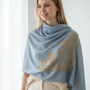 May include: A light blue shawl with gold embroidered detailing. The shawl is draped over the shoulders and features an ornate, corner design. The model is wearing a cream-colored top and pants.
