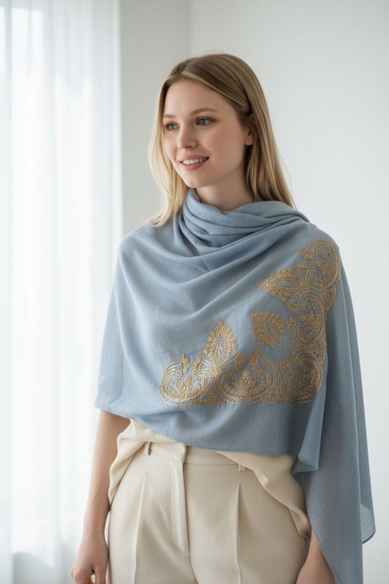 May include: A light blue shawl with gold embroidered detailing. The shawl is draped over the shoulders and features an ornate, corner design. The model is wearing a cream-colored top and pants.