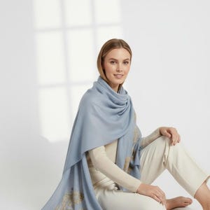 May include: A light blue scarf with gold embroidered detailing. The scarf is draped around the shoulders and arms. The person is wearing a cream-colored long-sleeve shirt and white pants. The scarf is a soft, flowing fabric.
