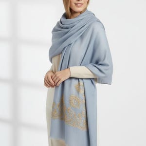 May include: A light blue scarf with gold embroidered paisley design. The scarf is draped over a cream-colored top and pants. The scarf has a soft, flowing appearance, suitable for cooler weather.