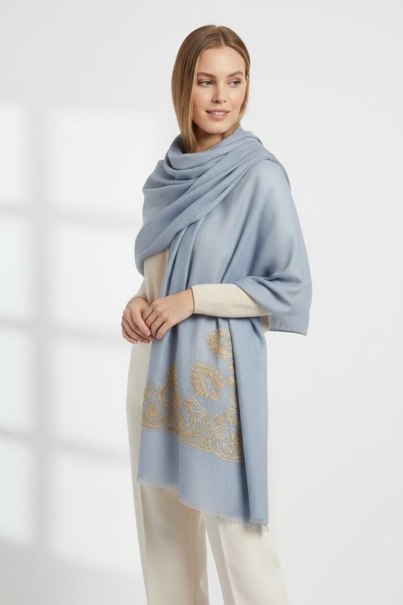 May include: A light blue scarf with gold embroidered paisley design. The scarf is draped over a cream-colored top and pants. The scarf has a soft, flowing appearance, suitable for cooler weather.