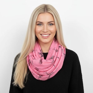 Puede incluir: Bufanda rosa infinita con un estampado de jirafas negras. La bufanda está envuelta alrededor del cuello y los hombros. La persona de la imagen lleva un jersey negro. El fondo es blanco.