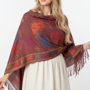 May include: A maroon shawl featuring a repeating pattern of elephants in various colors including orange, blue, and red. The shawl has a decorative border with smaller elephant motifs and long fringe trim along the edges, draped over a white dress.
