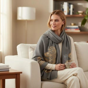 May include: A woman wearing a gray scarf with gold floral designs, seated on a white sofa. The scarf is draped around the neck and shoulders. She is holding a cup. A wooden side table with books is in the foreground.