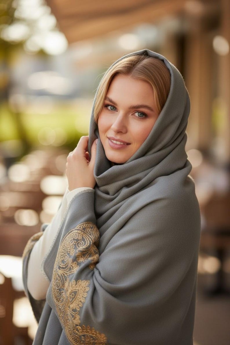 May include: A woman wearing a gray shawl with gold embroidery on the sleeves. The shawl is draped over the head and shoulders. The woman is smiling and looking at the camera.