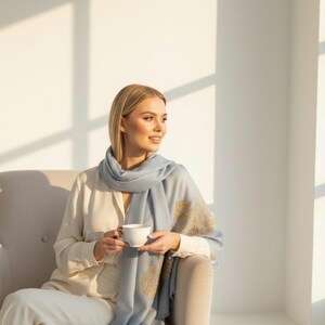 May include: A woman wearing a light blue scarf with gold embroidery, holding a white teacup. The scarf is draped over a cream-colored blouse and pants. The woman is seated in a beige armchair in a sunlit room.