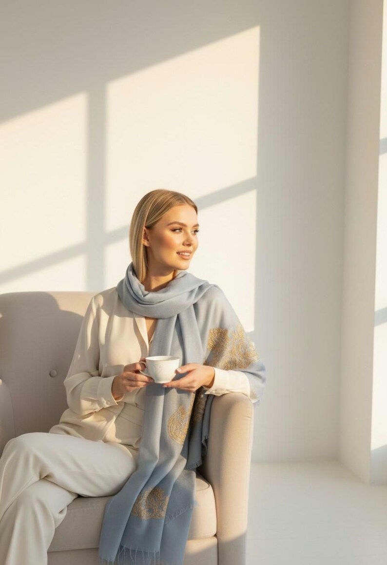 May include: A woman wearing a light blue scarf with gold embroidery, holding a white teacup. The scarf is draped over a cream-colored blouse and pants. The woman is seated in a beige armchair in a sunlit room.