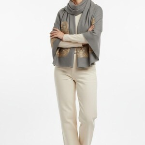 May include: A woman wearing a gray scarf with gold-colored decorative patterns, a cream-colored long-sleeve shirt, and cream-colored pants. The scarf is draped around the neck and over the shoulders. The pants are straight-legged.