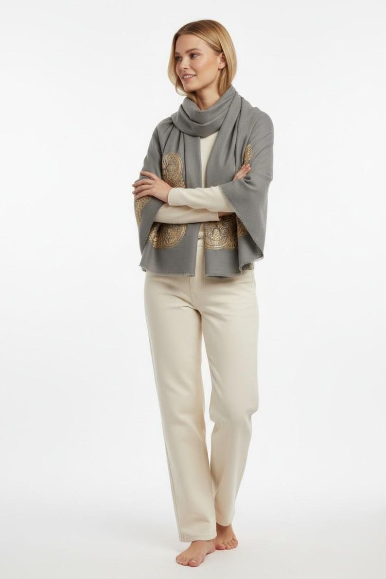 May include: A woman wearing a gray scarf with gold-colored decorative patterns, a cream-colored long-sleeve shirt, and cream-colored pants. The scarf is draped around the neck and over the shoulders. The pants are straight-legged.