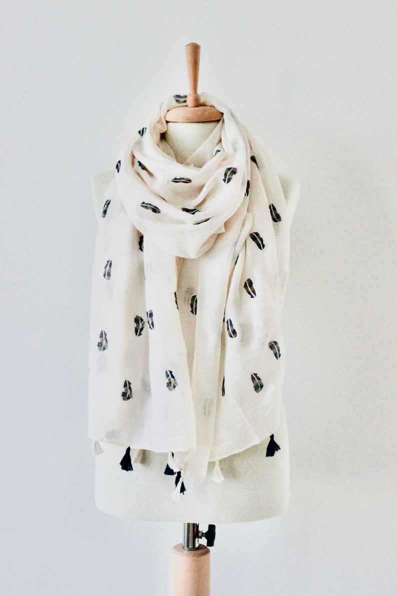 Peut inclure: Une &eacute;charpe blanche avec des motifs en forme de feuilles noires et brunes et des glands. L'&eacute;charpe est drap&eacute; sur un mannequin blanc.