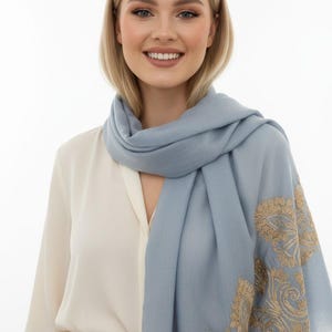 May include: A light blue scarf with intricate gold embroidery on one end. The scarf is draped around the neck and shoulders. The scarf is made of a soft material and has a fringed edge. The person is wearing a white blouse.