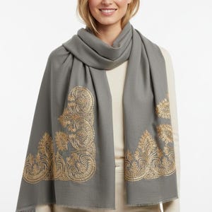May include: A gray scarf with gold embroidered paisley designs. The scarf is draped around the neck and shoulders. The scarf has a fringed edge.