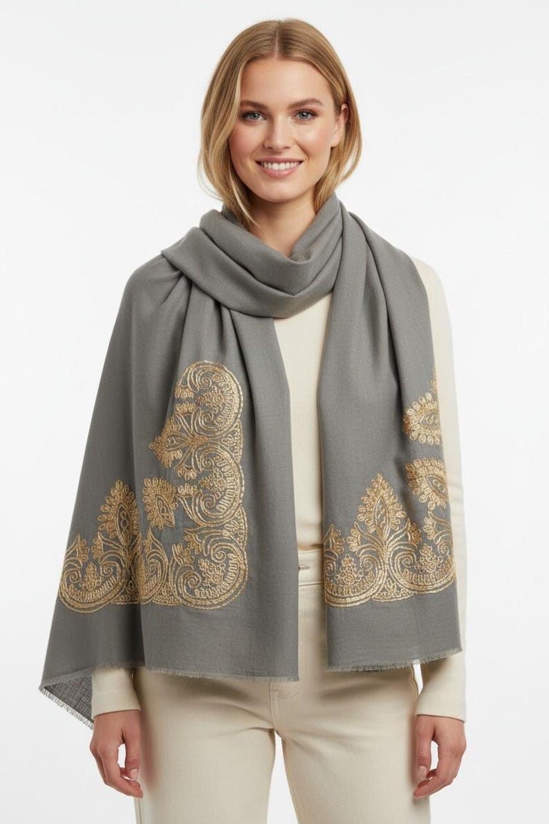 May include: A gray scarf with gold embroidered paisley designs. The scarf is draped around the neck and shoulders. The scarf has a fringed edge.