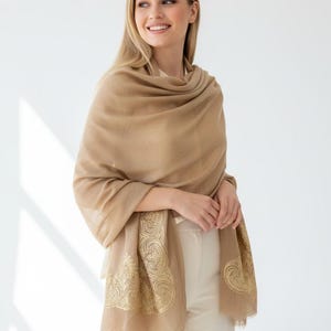 May include: A tan-colored shawl with a gold paisley design on the edges. The shawl is draped over the shoulders. The model is wearing a cream-colored top and pants. The background is white.