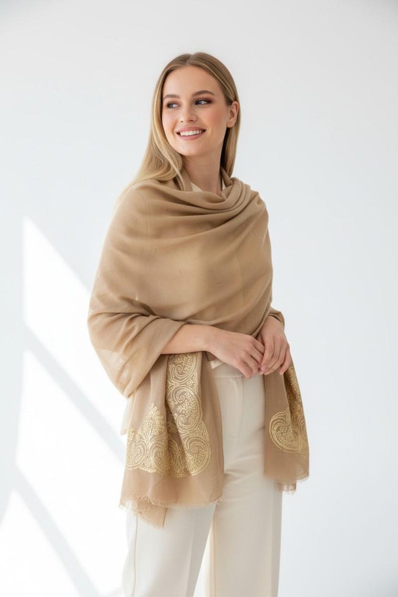 May include: A tan-colored shawl with a gold paisley design on the edges. The shawl is draped over the shoulders. The model is wearing a cream-colored top and pants. The background is white.