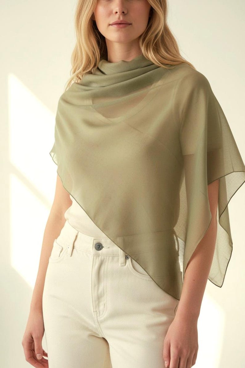 May include: A sheer, olive green scarf draped over the shoulders. The scarf has a flowing, lightweight appearance. The model is wearing a white top and cream-colored jeans. The scarf's edges are subtly outlined.