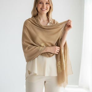 May include: A tan-colored shawl with gold embroidered floral designs on the edges. The shawl is draped over a cream-colored blouse and pants. The woman is smiling.