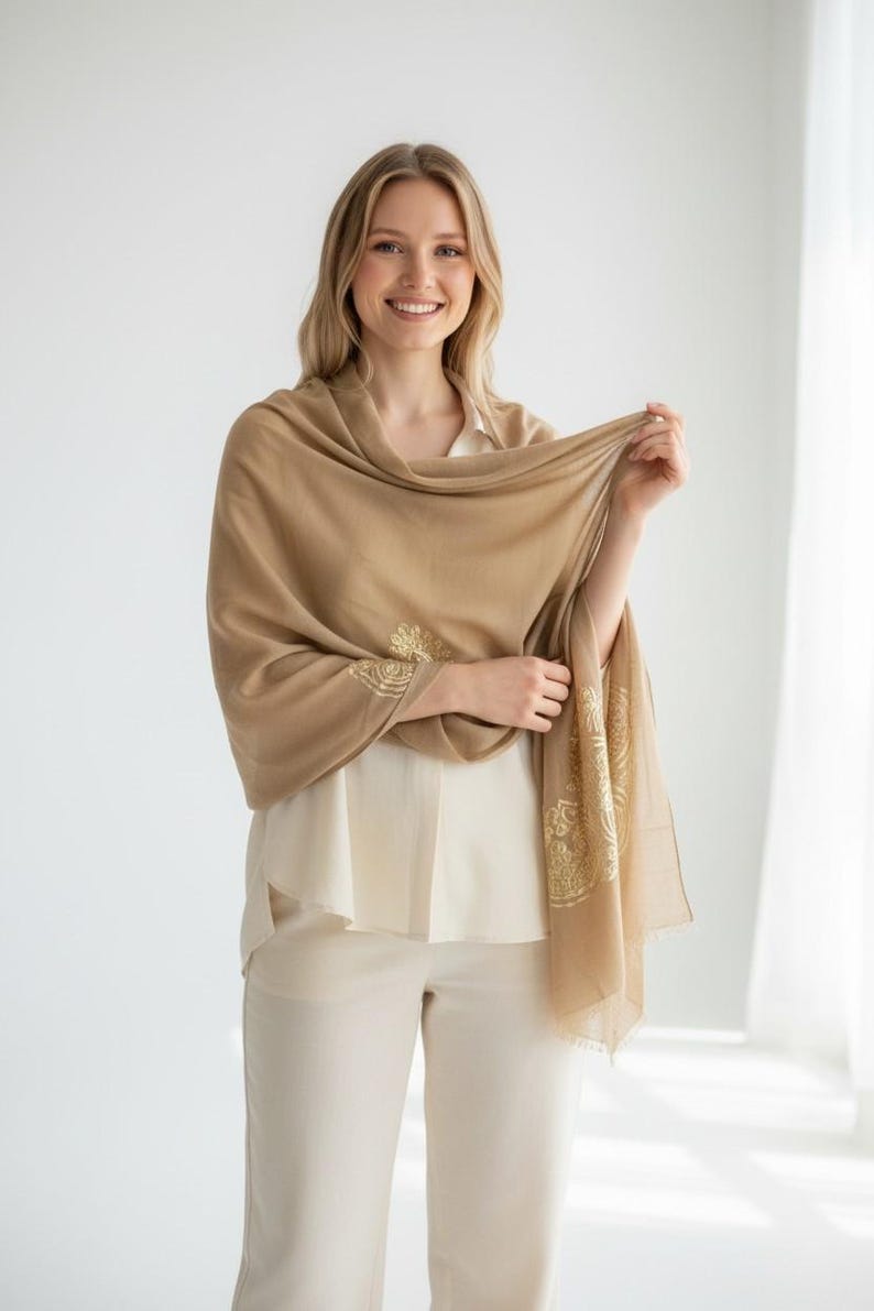 May include: A tan-colored shawl with gold embroidered floral designs on the edges. The shawl is draped over a cream-colored blouse and pants. The woman is smiling.