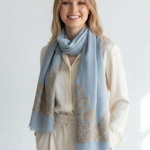 May include: A light blue scarf with gold embroidered paisley designs. The scarf is draped around the neck and shoulders. The scarf is made of a soft material and has a fringed edge. The model is wearing a cream-colored blouse and pants.