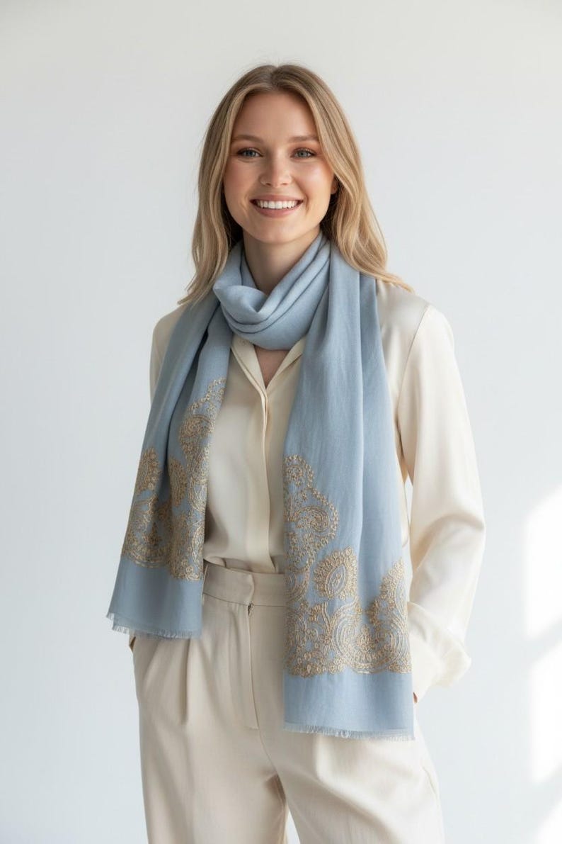 May include: A light blue scarf with gold embroidered paisley designs. The scarf is draped around the neck and shoulders. The scarf is made of a soft material and has a fringed edge. The model is wearing a cream-colored blouse and pants.