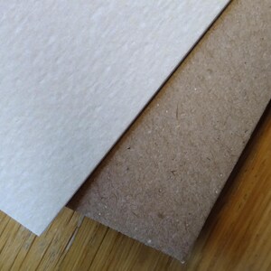 May include: Two pieces of paper, one white and one brown, are placed on top of a wooden surface. The white paper has a textured surface, while the brown paper has a rougher texture.