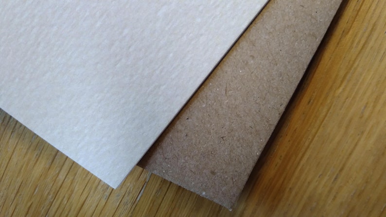 May include: Two pieces of paper, one white and one brown, are placed on top of a wooden surface. The white paper has a textured surface, while the brown paper has a rougher texture.