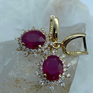 Könnte beinhalten: Goldene Ohrringe mit einem roten Rubin-Edelstein, der von einem Heiligenschein aus kleineren weißen Edelsteinen umgeben ist.