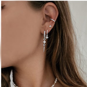May include: A delicate pearl necklace with gold accents. The necklace is made of small, white pearls and is worn by a person with long brown hair. The person is also wearing several silver earrings, including a hoop earring with a dangling charm.