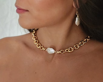 May include: A gold chain necklace with a large white pearl pendant. The necklace is a choker style and is worn by a person with dark hair.