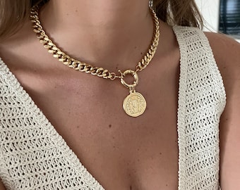 May include: A gold chain necklace with a large, round coin pendant. The coin has an intricate design and is attached to the chain with a large, gold ring.