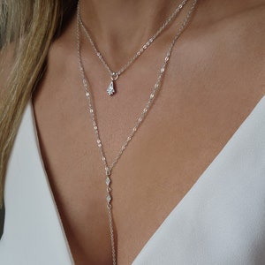 Pu&ograve; includere: Una collana argentata a due fili con un piccolo ciondolo a forma di lacrima. Il ciondolo ha un design geometrico ed &egrave; decorato con piccole pietre trasparenti. La collana &egrave; indossata da un manichino che indossa una camicetta bianca.