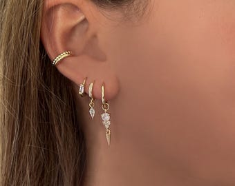May include: Gold hoop earrings with a dangling charm featuring a small triangle and three clear cubic zirconia stones.