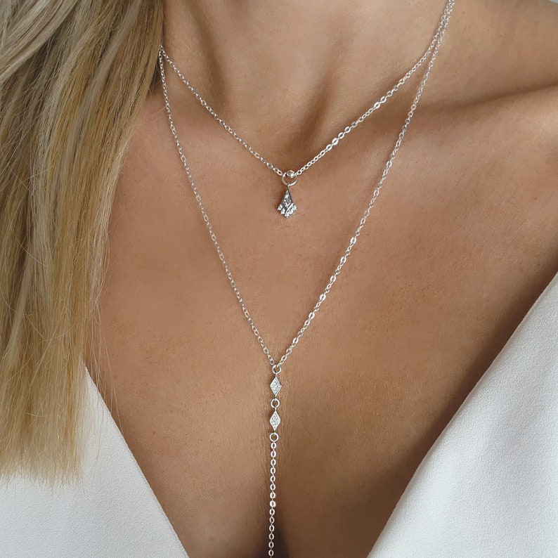 Pu&ograve; includere: Una collana argentata a due fili con un piccolo ciondolo a forma di diamante sulla catena pi&ugrave; corta e una catena pi&ugrave; lunga con tre ciondoli a forma di diamante.
