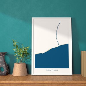 Op de afbeelding: Een witte ingelijste print met een abstract ontwerp in blauw en wit. De print heeft de tekst "SIDMOUTH DEVON" onderaan. De print staat op een houten plank met een vaas en een potplant.