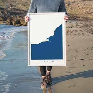Könnte beinhalten: Ein gerahmter Kunstdruck mit einem minimalistischen Design einer Küstenlandschaft in Weiß und Marineblau. Der Druck enthält das Wort "FOLKESTONE". Das Kunstwerk wird von einer Person an einem Sandstrand gehalten.