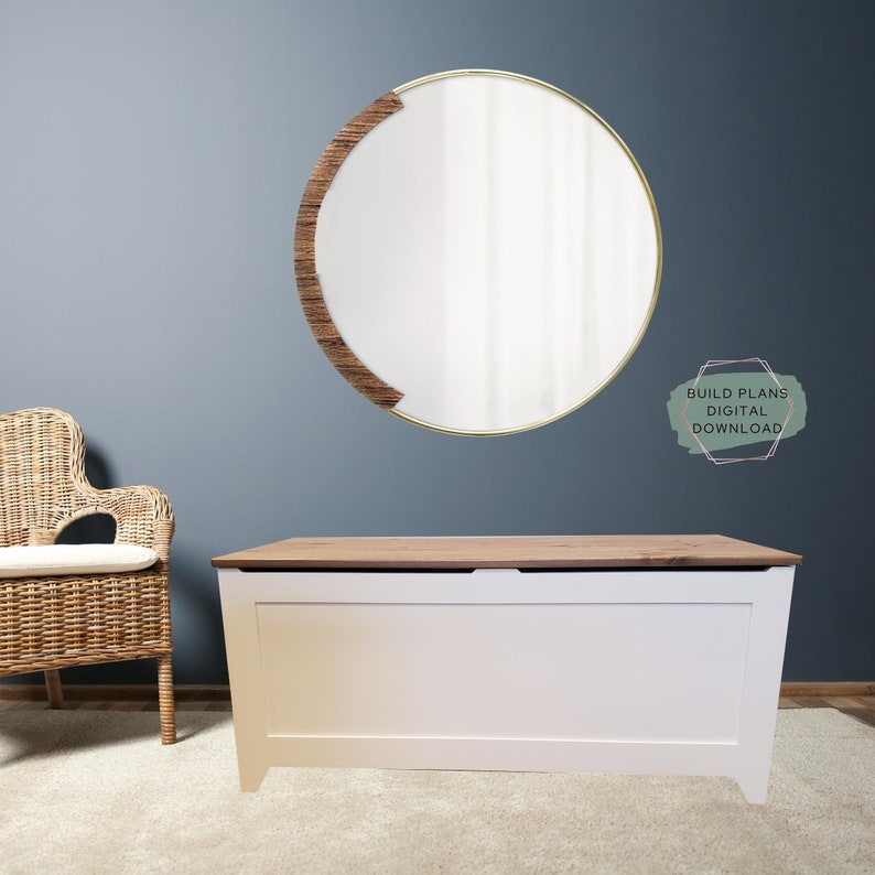 May include: White toy chest with a wood top lid. The chest is sitting on a light beige rug in front of a blue wall. A round mirror with a wood frame is hanging on the wall. The text "BUILD PLANS DIGITAL DOWNLOAD" is written in a green circle on the wall.