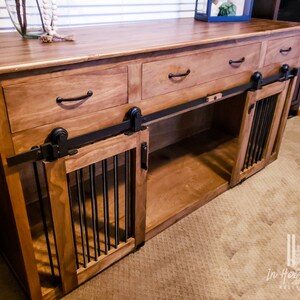 May include: A wooden dog crate with two sliding doors and three drawers. The crate is made of light brown wood and has black metal accents. The doors have a black metal grate and the drawers have black metal handles.
