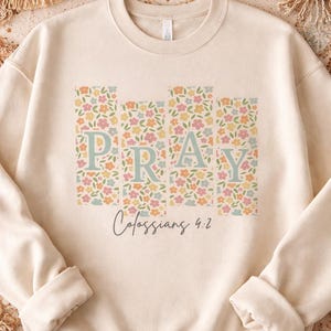 Pode incluir: Camisola creme com a palavra "PRAY" em letras azuis pastel, cada uma sobre um fundo floral. O texto "Colossenses 4:2" está escrito abaixo em uma fonte cursiva. A camisola tem gola redonda e mangas compridas.